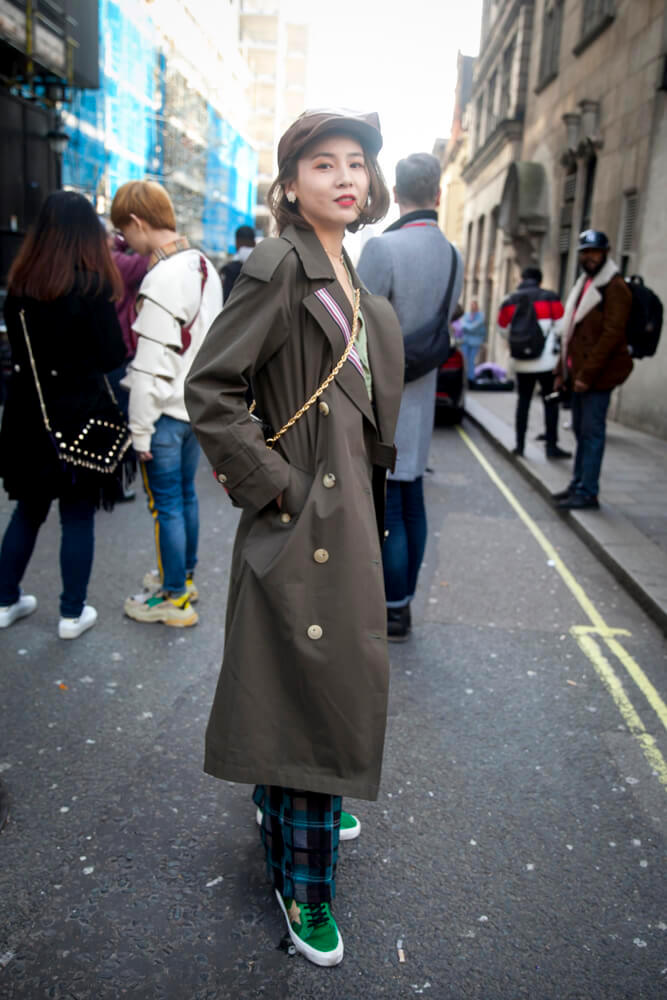 LONDON - FEBRUARY 17, 2019: Stylish attendees gathering outside 180 The Strand for London Fashion Week. The girl wears a beige trench coat and checkered wide trousers and a cap.