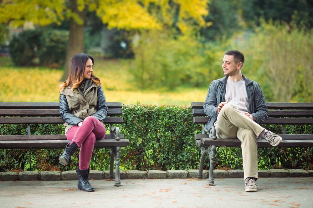 Man and woman smiling at each other across two different benches in the park