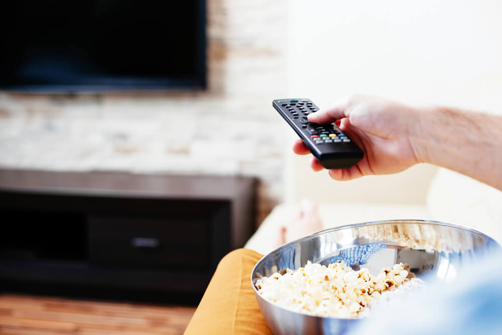 Hand holding remote pointing at TV, with popcorn on lap