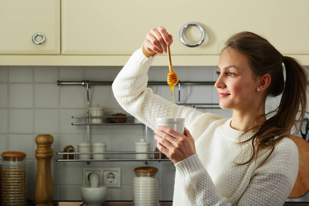 Woman adding honey to mug in kitchen